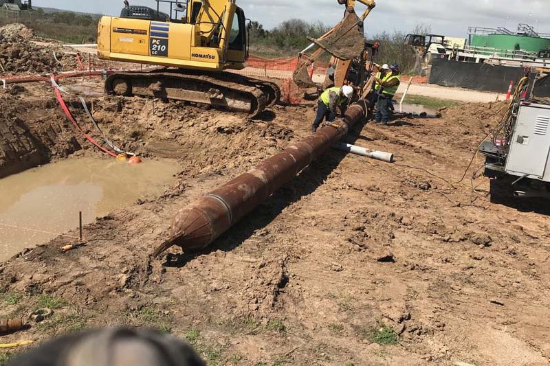 Excavator lowering a length of steel casing pipe into an open excavation on a jobsite